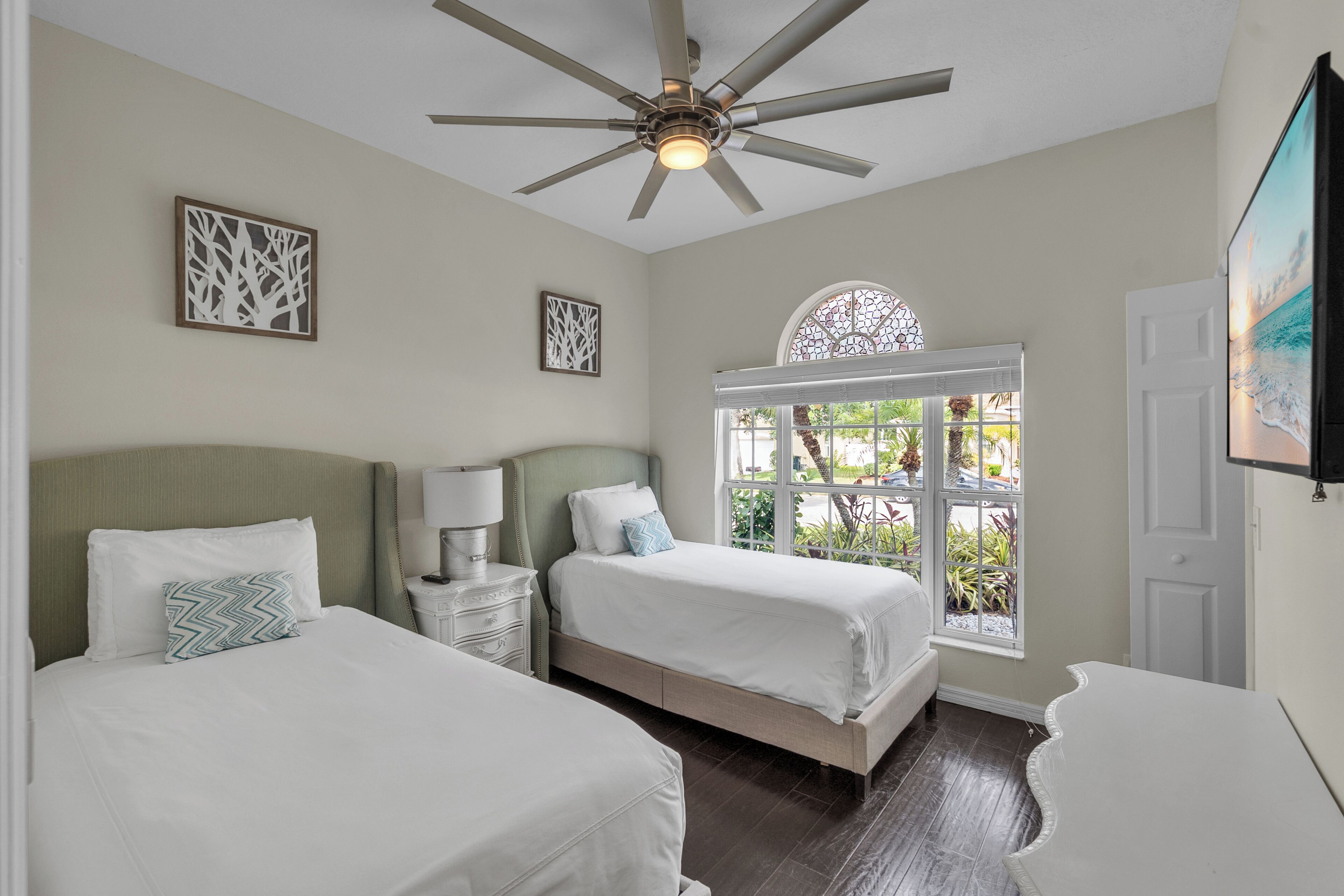 A hotel room with two beds, a ceiling fan, a window with a view, and artwork on the walls.