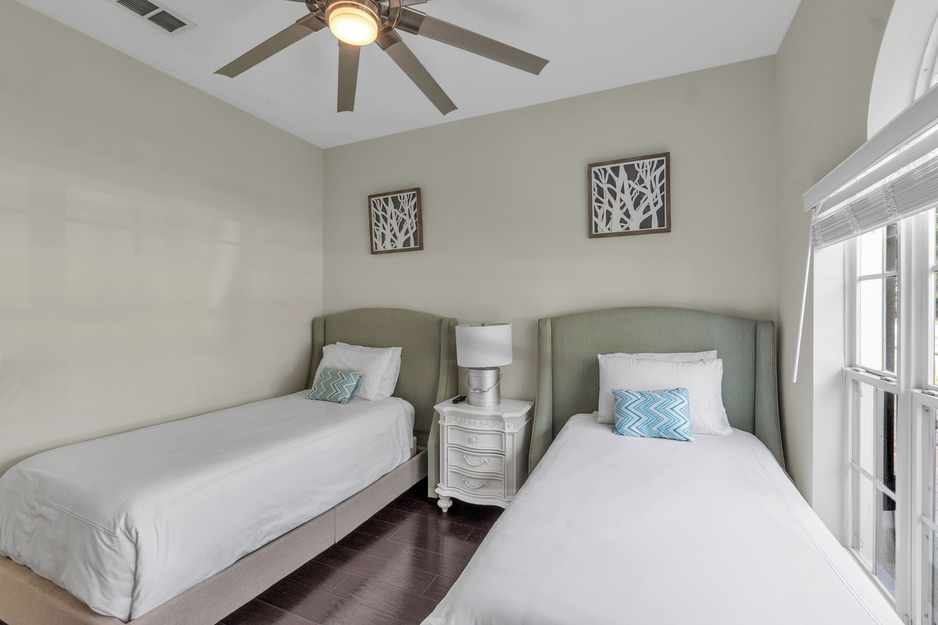 Une chambre d’hôtel avec deux lits simples, une table de chevet avec une lampe, des œuvres d’art décoratives au mur, un ventilateur de plafond et une grande fenêtre.