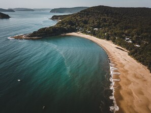 Vista aérea de una zona costera con playa arenosa, aguas turquesas cristalinas y densas colinas verdes al fondo.