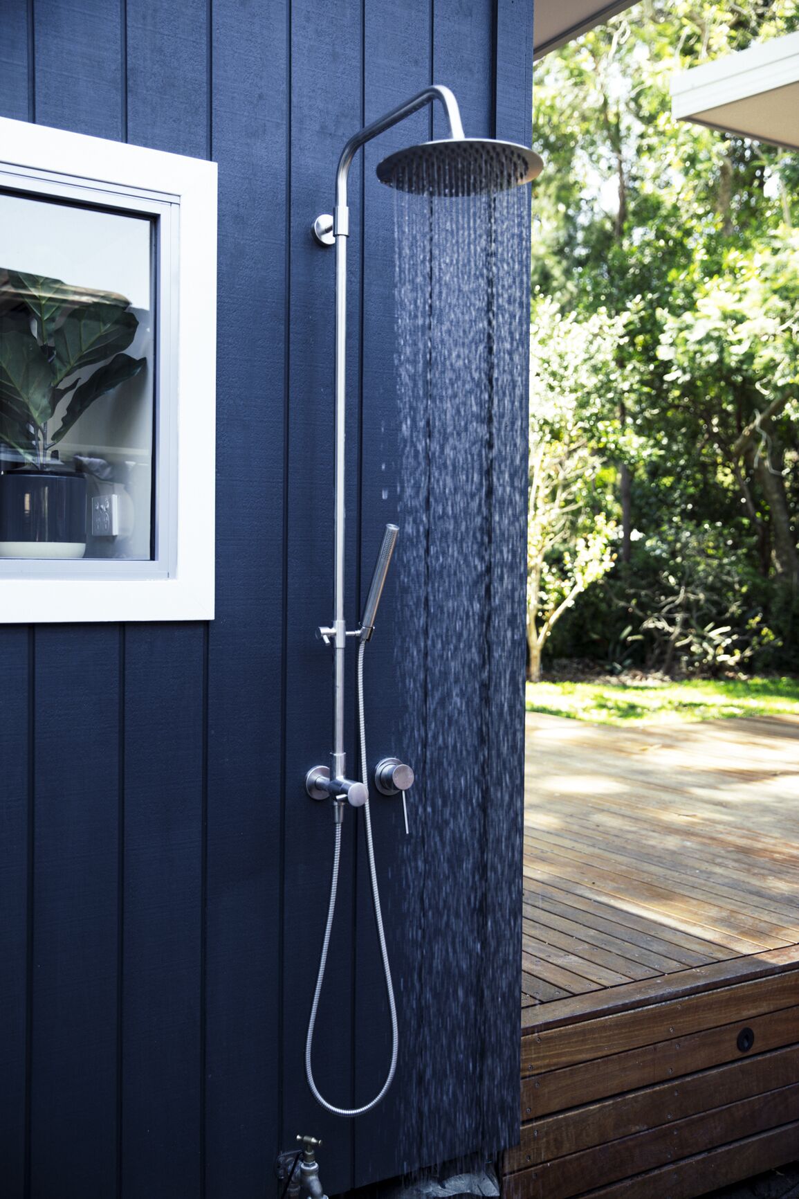 Une douche d’extérieur moderne, dotée d’un pommeau de douche à effet pluie, d’un pommeau de douche à main et d’un flexible à main, le tout fixé sur un mur en bois foncé.