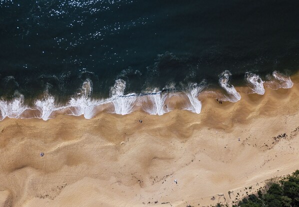 Vista aérea de una playa con olas rompiendo en la orilla, algunas personas dispersas a lo largo de la playa y un cielo despejado.