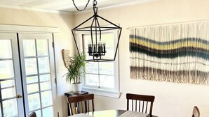 A dining area with a round table, six wooden chairs, a central light fixture, a potted plant, and a wall-mounted macrame piece. - The Farmhouse @ Four Lanterns Winery (Paso Robles)