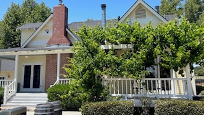 A two-story house with a brick chimney, a white porch, and a garden with green foliage. - The Farmhouse @ Four Lanterns Winery (Paso Robles)