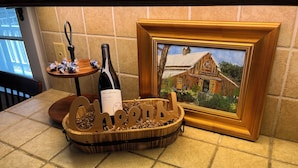 A wooden tray with a bottle of wine, a small table with decorative items, and a framed painting on a tiled wall. - The Farmhouse @ Four Lanterns Winery (Paso Robles)