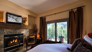 Ein Schlafzimmer mit Kamin aus Stein, einem großen Bett mit Zebra-Kissen, einem Fernseher auf einer Ablage und Blick auf Bäume durch ein Fenster.