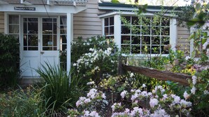 Une petite maison avec une porte et des fenêtres blanches, entourée d’une végétation luxuriante et de plantes en fleurs.