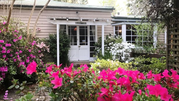 Une maison dotée d’un porche abrité, d’une enseigne indiquant « Hampden Cottage », et d’un jardin où poussent des fleurs roses et blanches.
