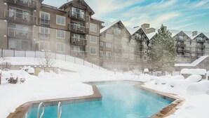 Une piscine extérieure entourée de neige, avec une eau d’un bleu limpide, bordée d’immeubles à plusieurs étages dotés de balcons, le tout dans un paysage hivernal enneigé.