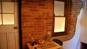Eine Ziegelwand mit einem Fenster, ein Holztisch mit einer Obstschale und ein Stuhl.