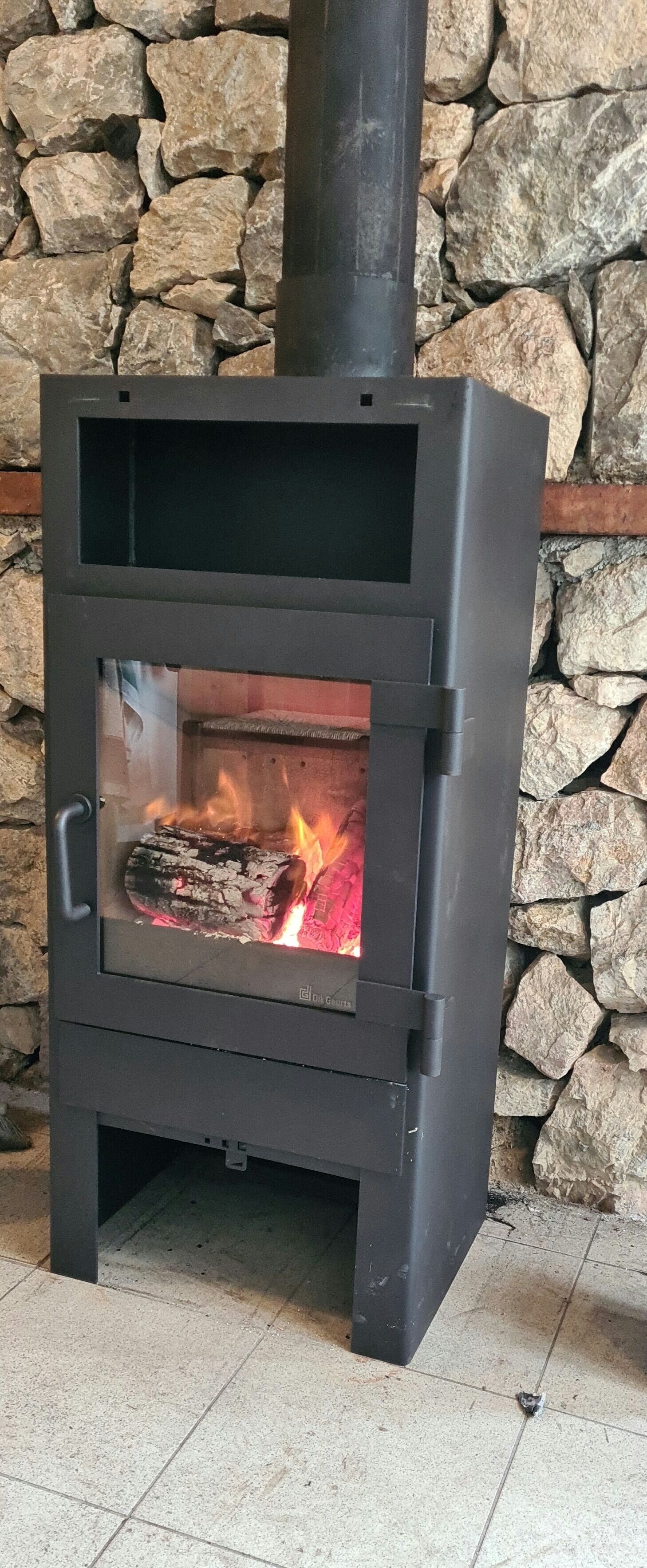 Una estufa de leña con puerta de cristal, frente a una pared de piedra.