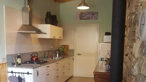 Keittiössä on valkoiset kaapit, liesi ja seinälle ripustettuja keittiövälineitä. Taustalla on kiviseinä ja ovi.
