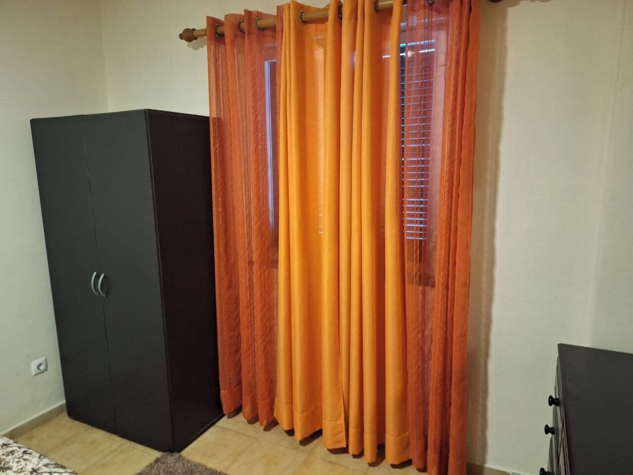 A room with a wardrobe, orange curtains, a window with blinds, and a small cabinet.