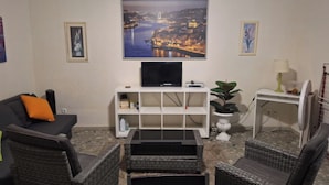A living room with a flat-screen TV on a white stand, wicker furniture, a small white table with a lamp, and a framed cityscape painting on the wall.