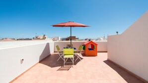 Eine Dachterrasse mit einem roten Sonnenschirm, zwei grünen Stühlen, einem kleinen Spielhäuschen und einem Tisch.