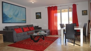 A living room with a grey sofa, red cushions, a coffee table, and a red rug. There is a dining area with a table and chairs, and a window with red curtains. - Discover the originality of CAP (P.M.R. access)7 pers (Sisco)