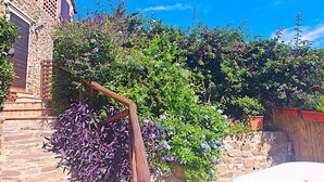 Un edificio in pietra con una scala in legno che conduce a una porta, circondato da vegetazione lussureggiante e piante fiorite.