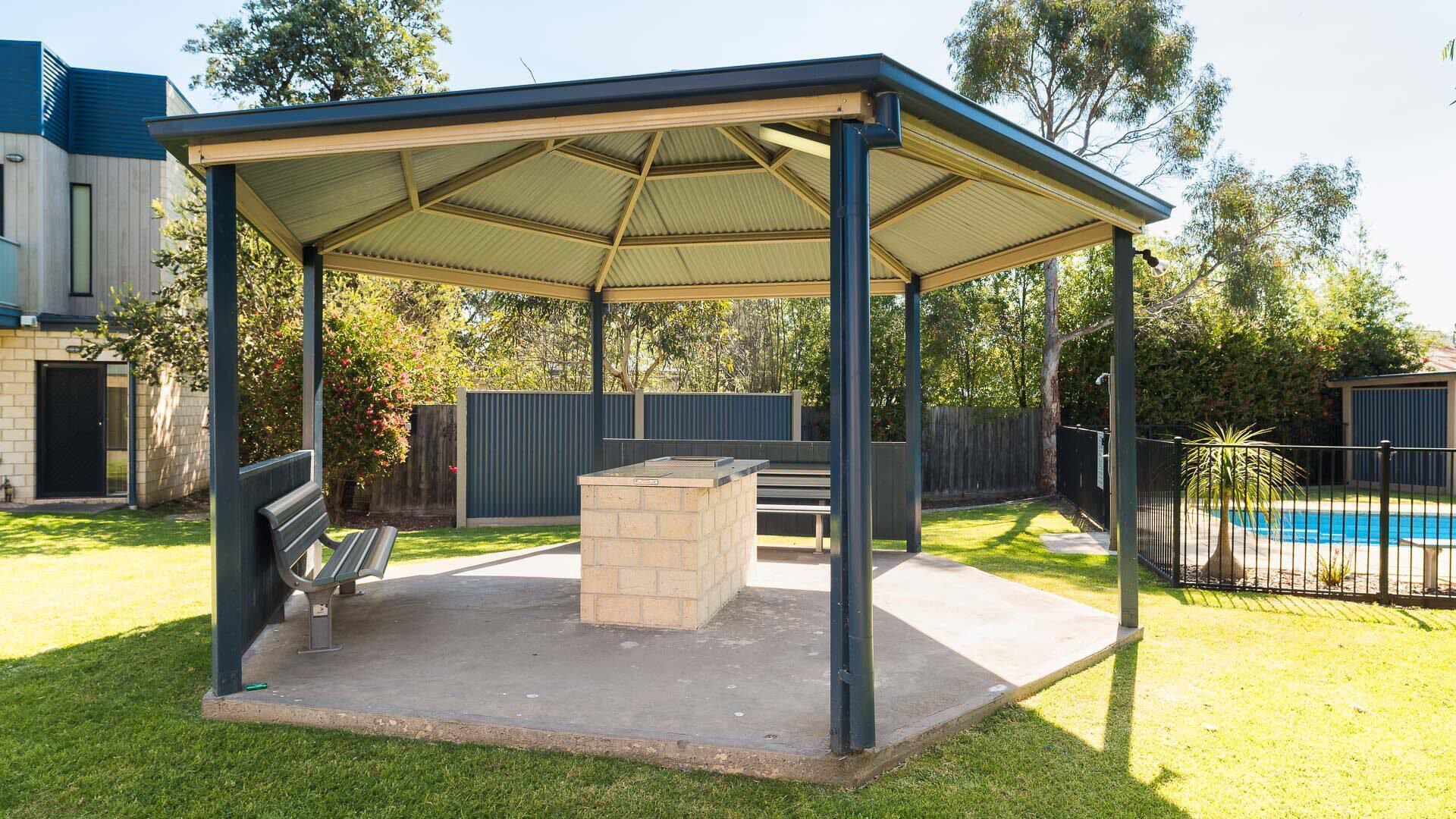 A covered outdoor area with a barbecue station, a bench, and a pool in the background.