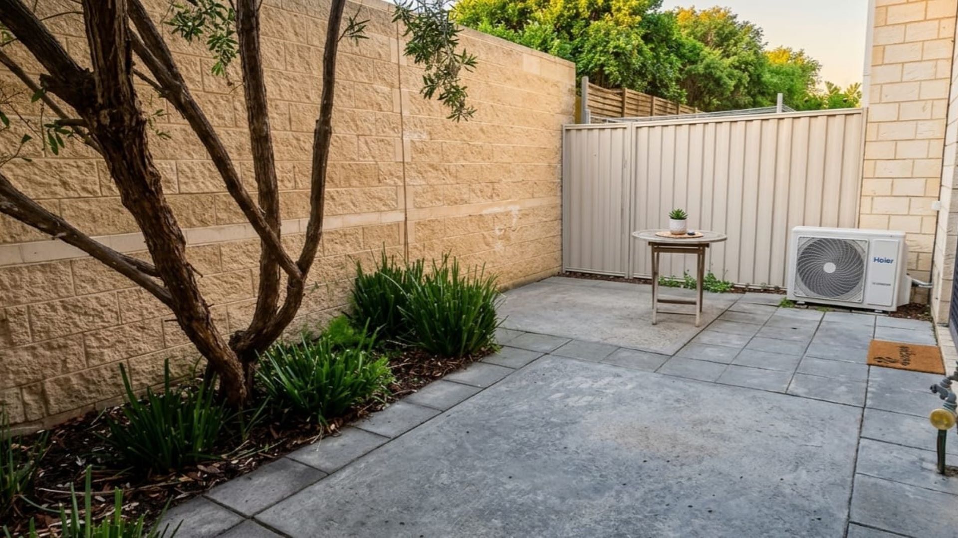 A paved patio with a small table and a fan, surrounded by a low stone wall and a tree.