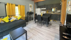 Un espacio habitable compacto con cama, una mesa de comedor con sillas, una kitchenette y una pequeña mesa con lámpara.