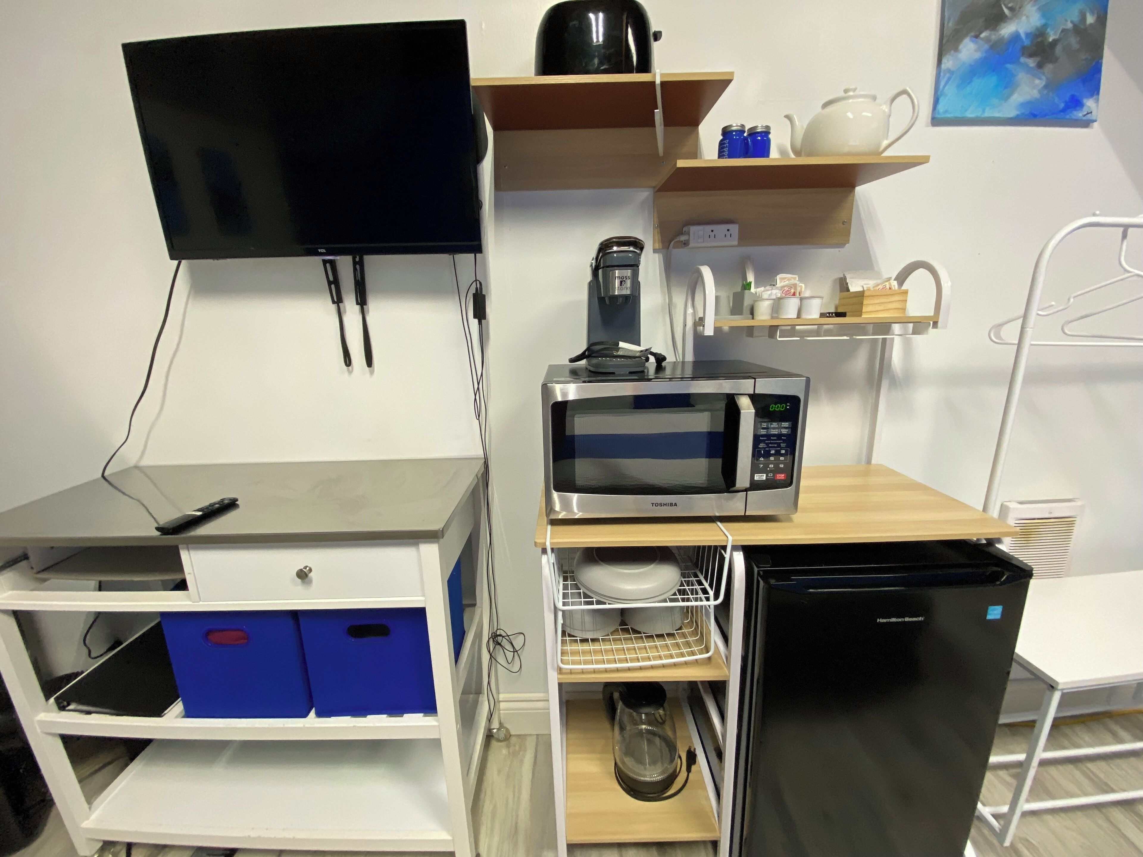 A compact kitchenette with a microwave, kettle, and coffee maker. There is a TV mounted on the wall, a small shelf with tea and condiments, and a refrigerator.