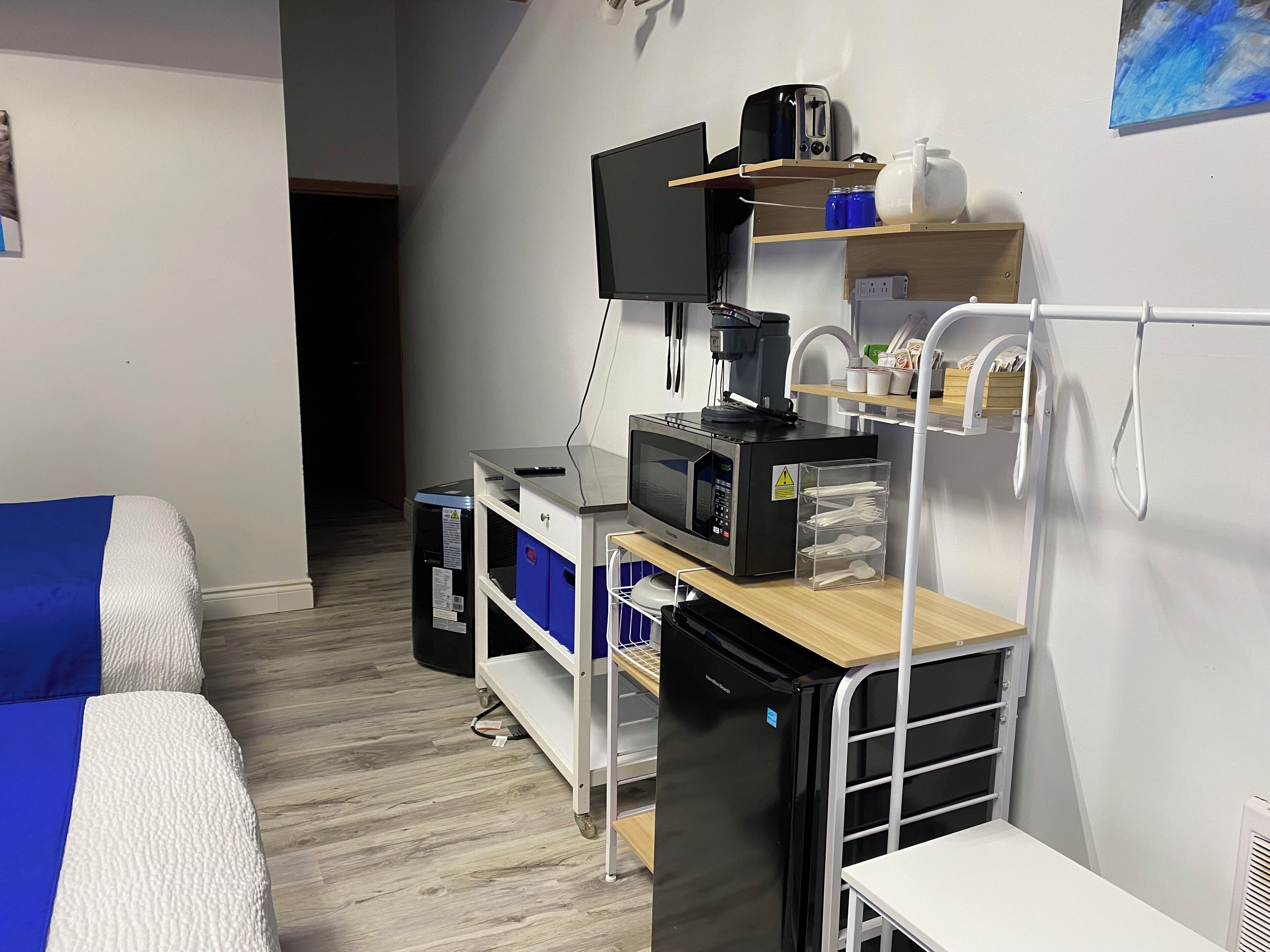 Quarto de hotel compacto com cama, escrivaninha com computador, micro-ondas e pequena área de cozinha.