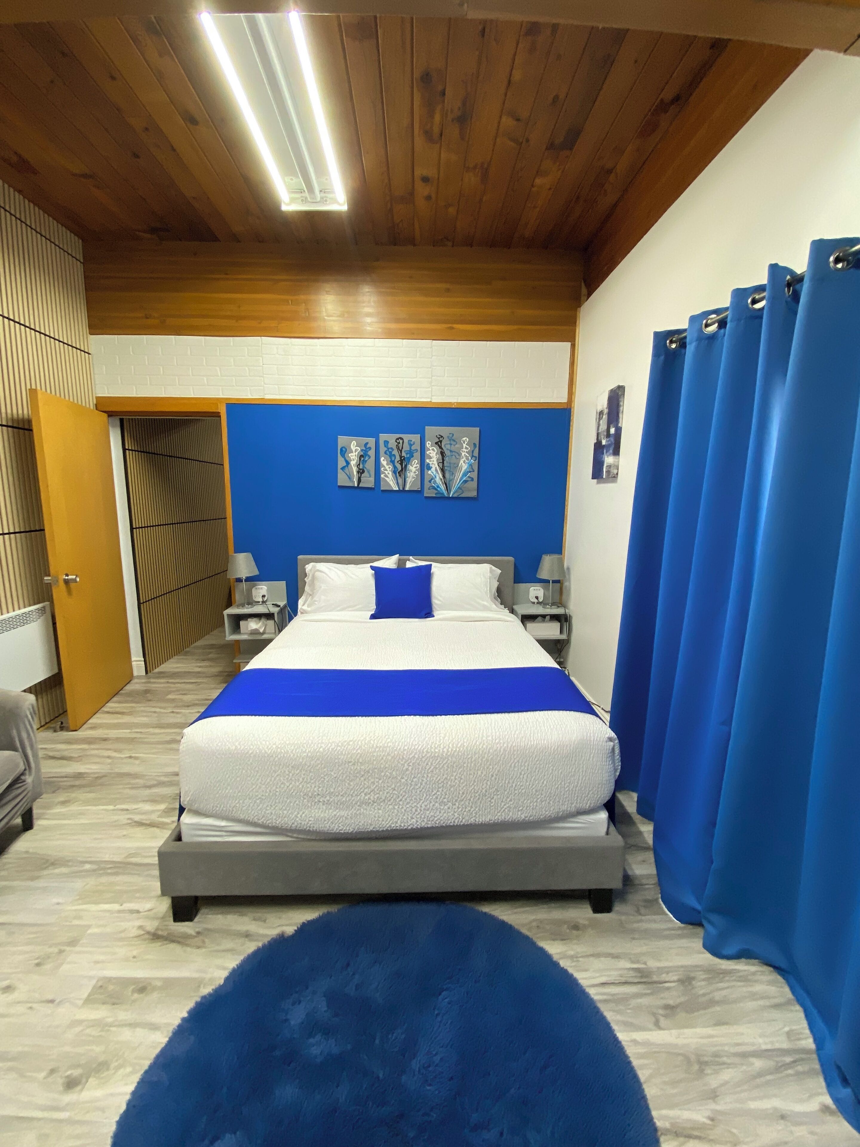 A bedroom with a bed, a blue rug, a wooden ceiling, and a blue curtain.