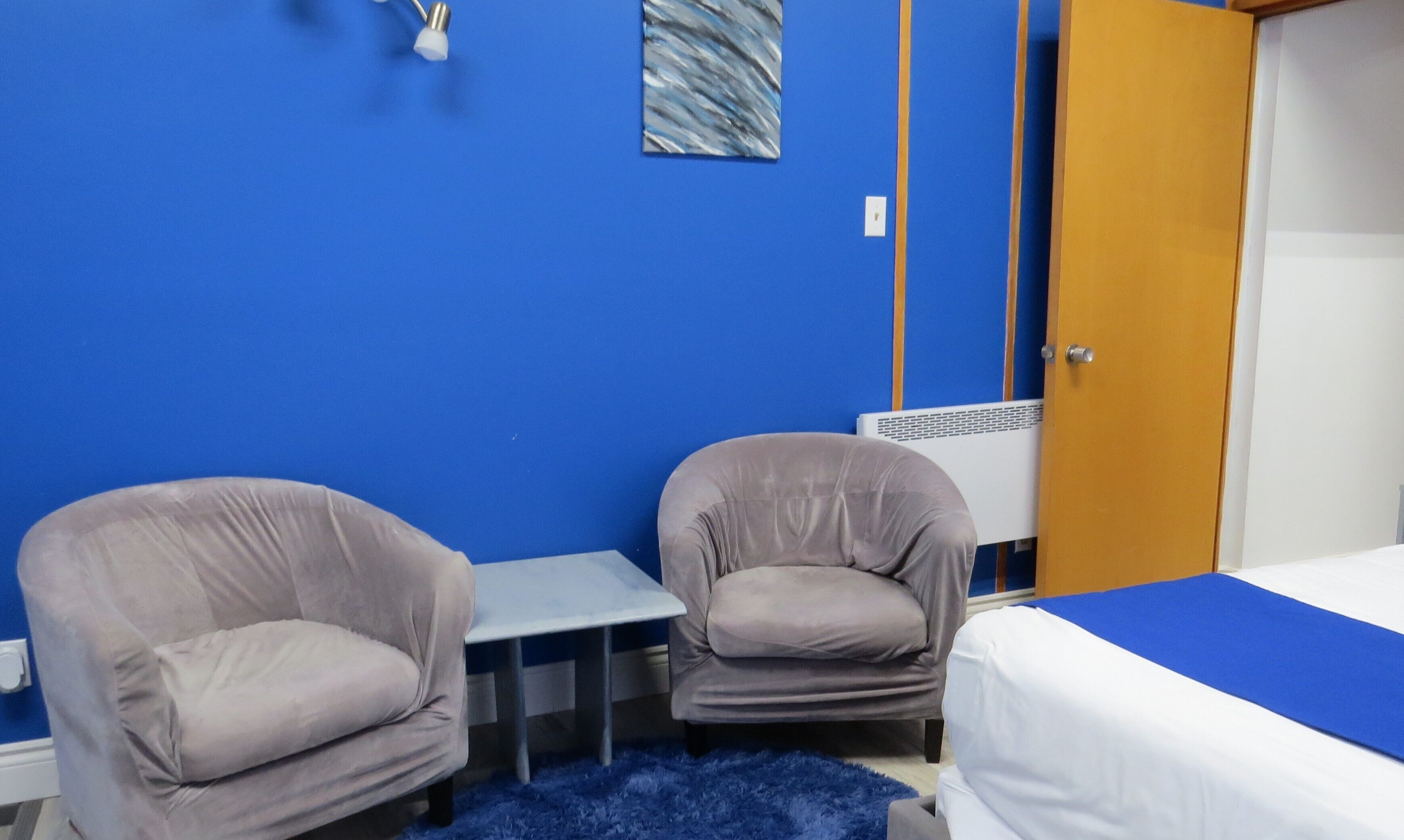 A room with two armchairs, a small table, a bed with a blue bedspread, a radiator, and a door.