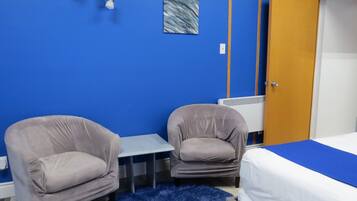 A room with two armchairs, a small table, a bed with a blue bedspread, a radiator, and a door.