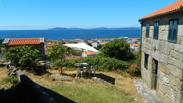 A stone building with a tiled roof, a garden with a table and chairs, and a view of the sea and distant buildings. - Little house with seaviews (Pedrafigueira)