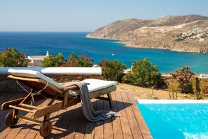 Une terrasse en bois avec une piscine, des chaises longues et une vue sur la mer et les collines.