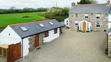 A stone house with a sloped roof and a white exterior, a gravel driveway, and a green field in the background.