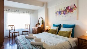 Ett hotellrum med en säng, två stolar, ett skrivbord, en lampa och en tavla med en stadsbild på väggen.