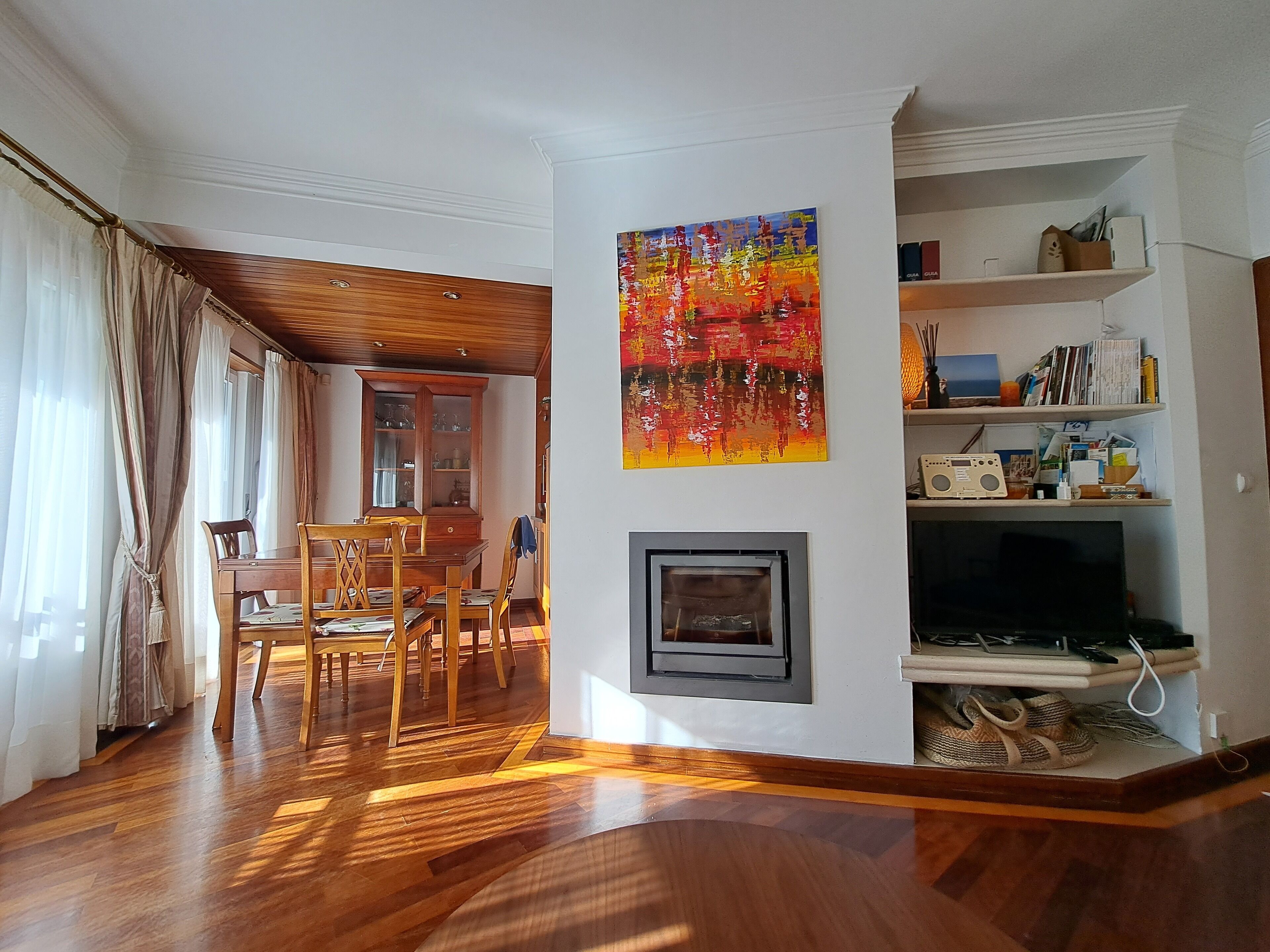 Un salon doté d’un plancher en bois, un coin repas avec une table et des chaises en bois, un meuble avec des portes en verre, une cheminée et une bibliothèque contenant divers objets.