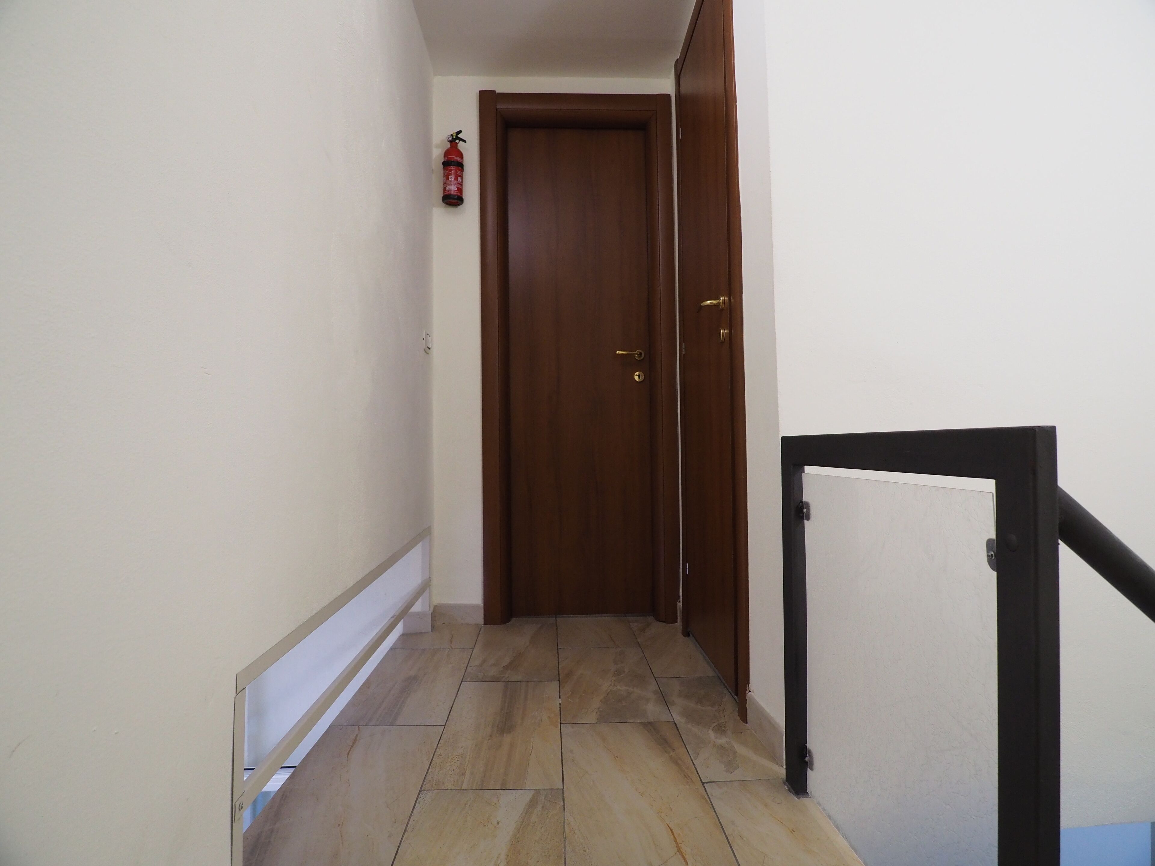 A narrow hallway with a wooden door at the end, tiled flooring, and a fire extinguisher mounted on the wall.