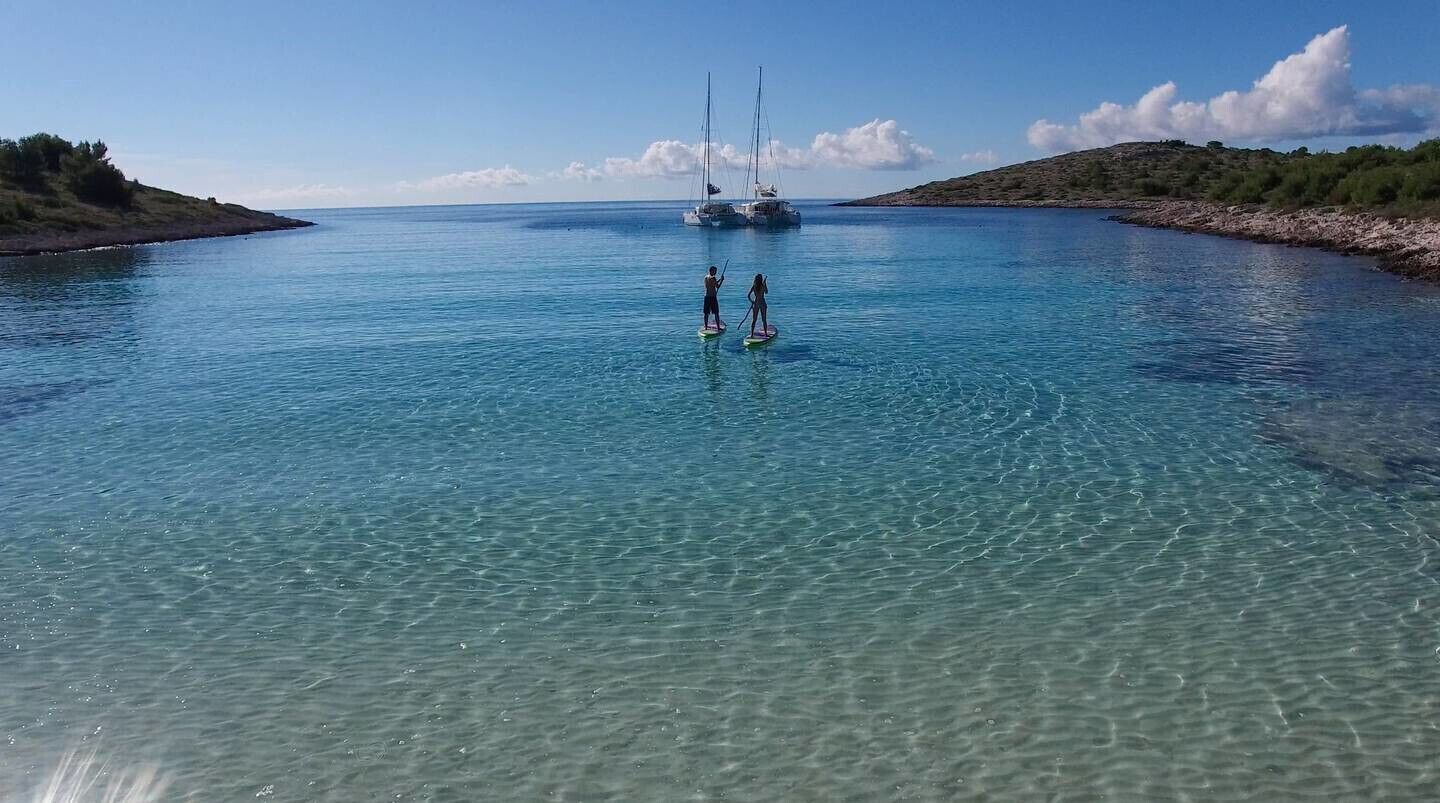 Klares Küstengewässer mit zwei Stand-Up-Paddlebrettern, einem entfernten Segelboot und felsigem Ufer.