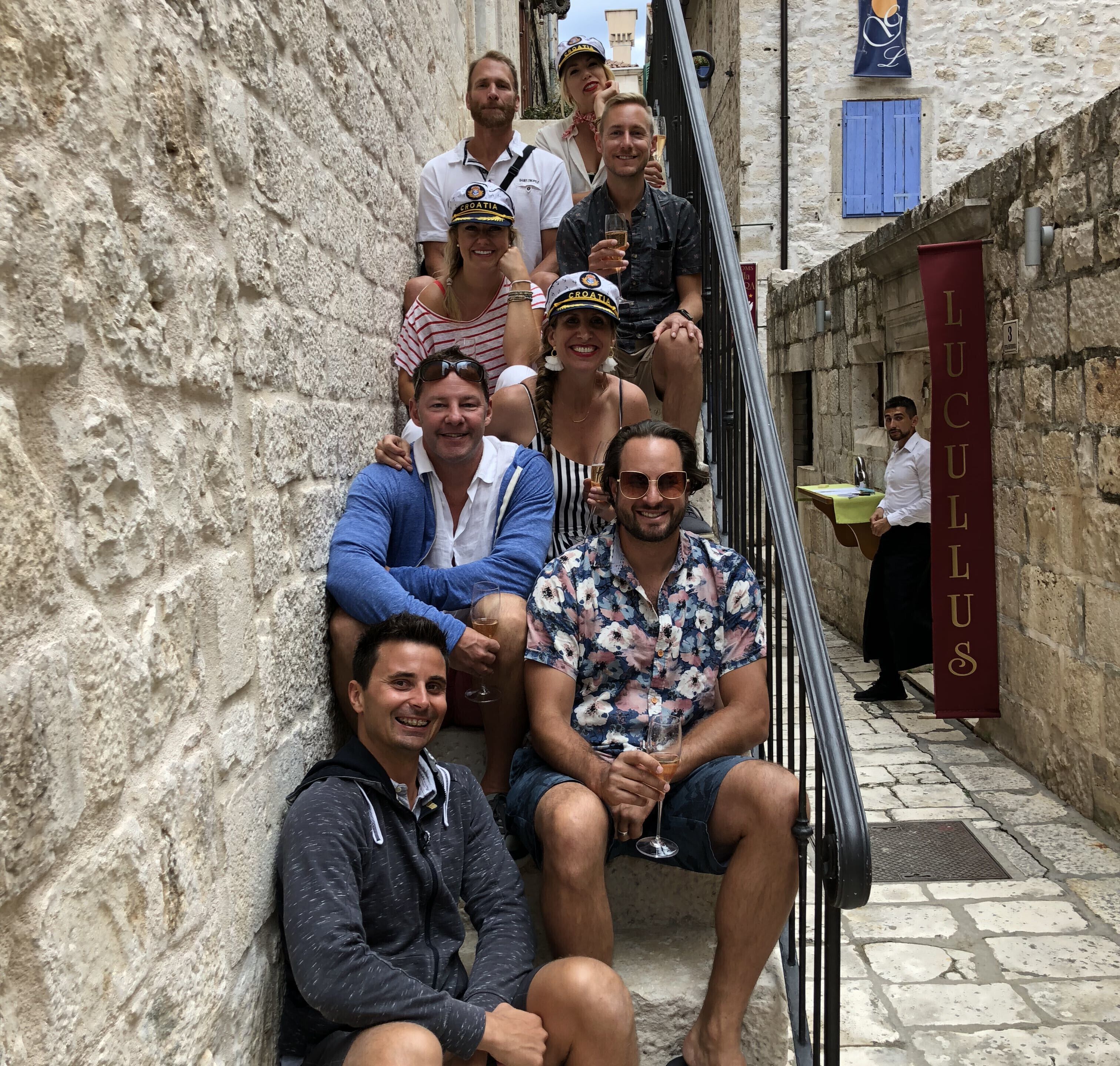 A group of people sitting on a stone staircase in a narrow alleyway with a sign reading "LUCULUS".