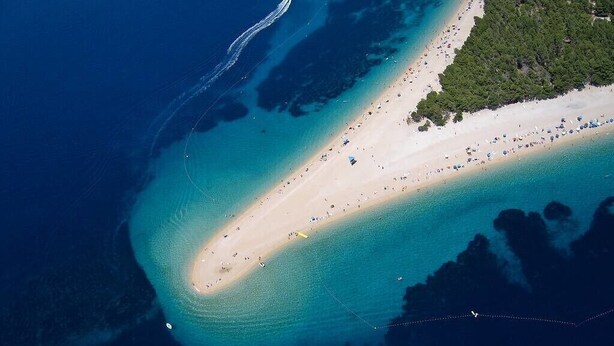 Vista aérea de una playa con aguas cristalinas, una orilla de arena y vegetación densa a la derecha.