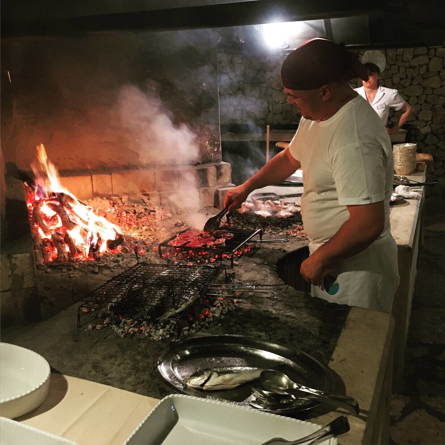 A chef grilling meat over an open flame in a rustic outdoor kitchen.