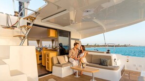 Interior de un yate con comedor, zona de estar con sofá y mesa, y vista al mar.