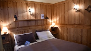 Una habitación con paneles de madera, una cama, dos lámparas de noche, una estantería con un libro y un aplique en la pared.