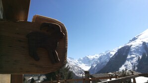 Un letrero de madera con una figura de animal tallada, una cerca de madera y un paisaje montañoso con picos nevados al fondo.