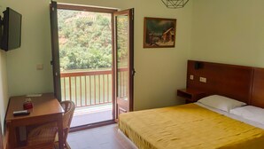 Un dormitorio con cama, escritorio, silla, un cuadro y vista a un río y árboles por una puerta abierta.