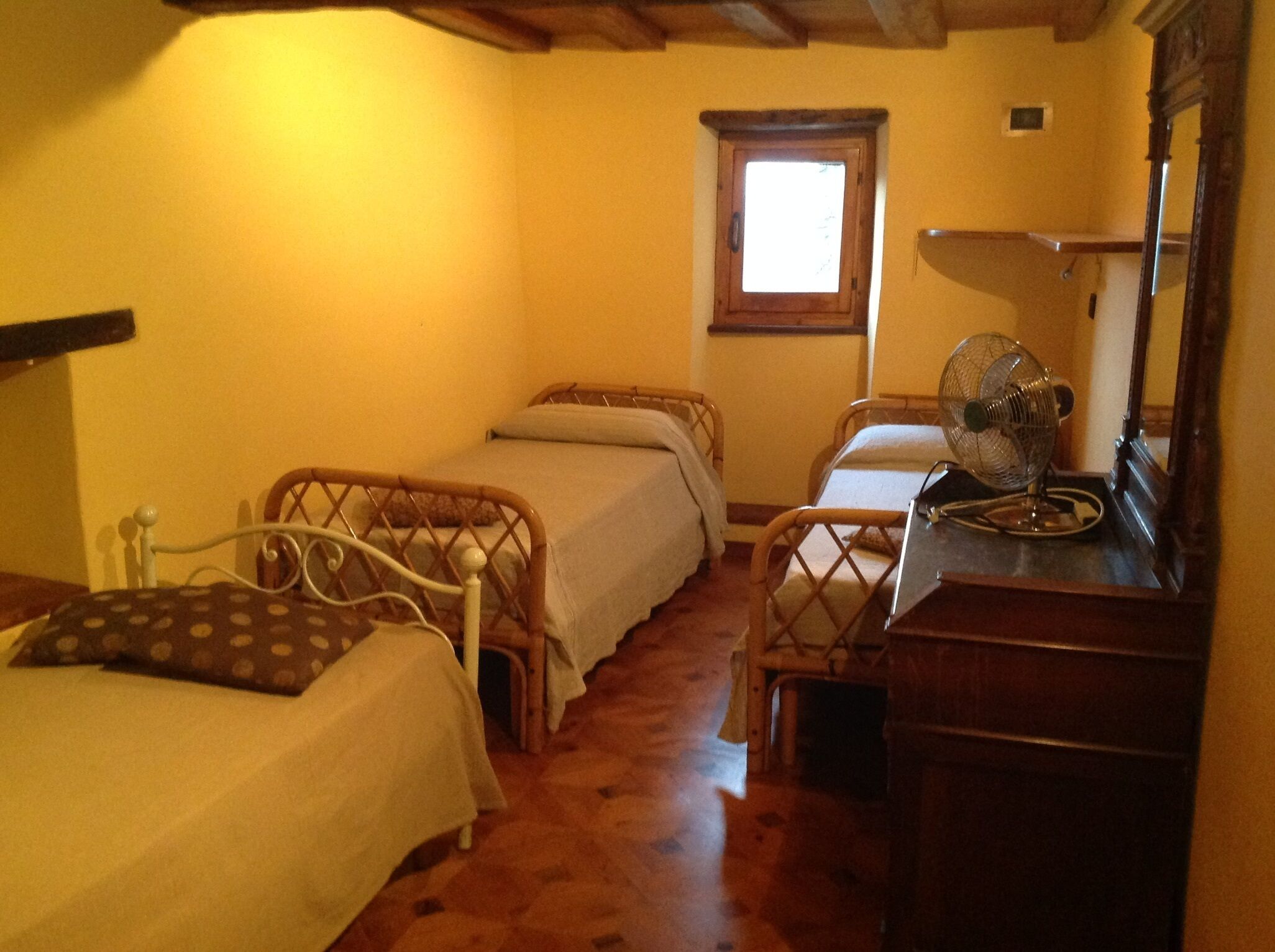 A small, cozy room with two single beds, a wooden floor, a window with a view, a wooden dresser, and a fan.