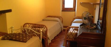 A small, cozy room with two single beds, a wooden floor, a window with a view, a wooden dresser, and a fan.