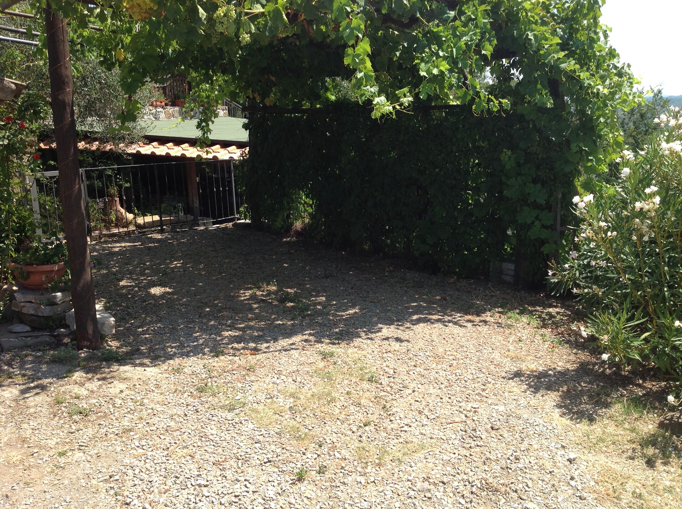 A gravel courtyard with a pergola covered in climbing plants, a fenced area with a tiled roof, and a stone wall.