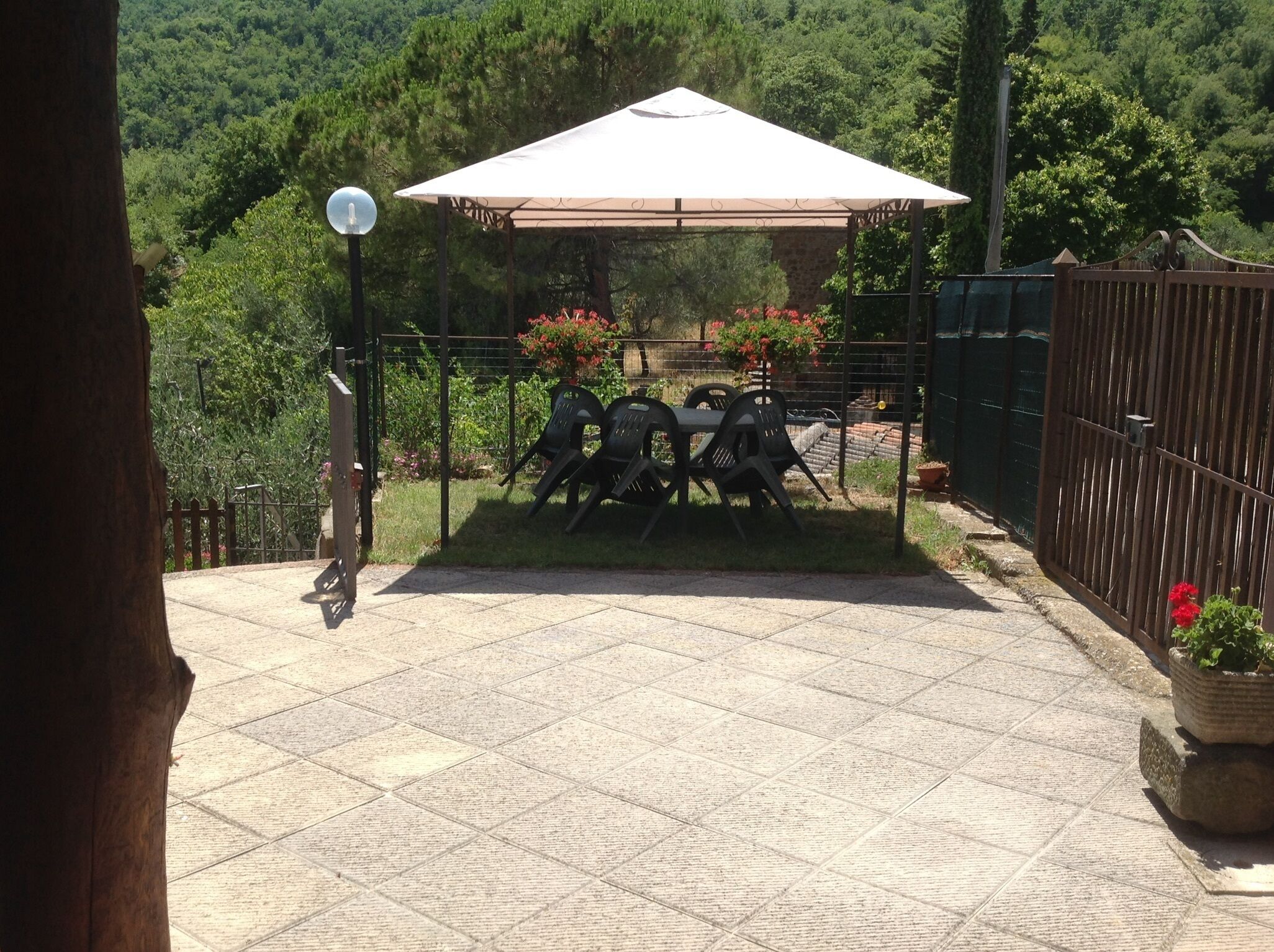A patio area with a table and chairs under a pergola, surrounded by a fence and greenery.
