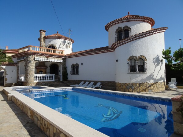 A poolside view of a building with a swimming pool, stone walls, and a tiled roof.