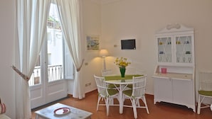 A dining area with a round table, white chairs, a vase with yellow flowers, a cabinet with glass doors, and a window with white curtains. - Stylish Apartment - Minori (Minori)