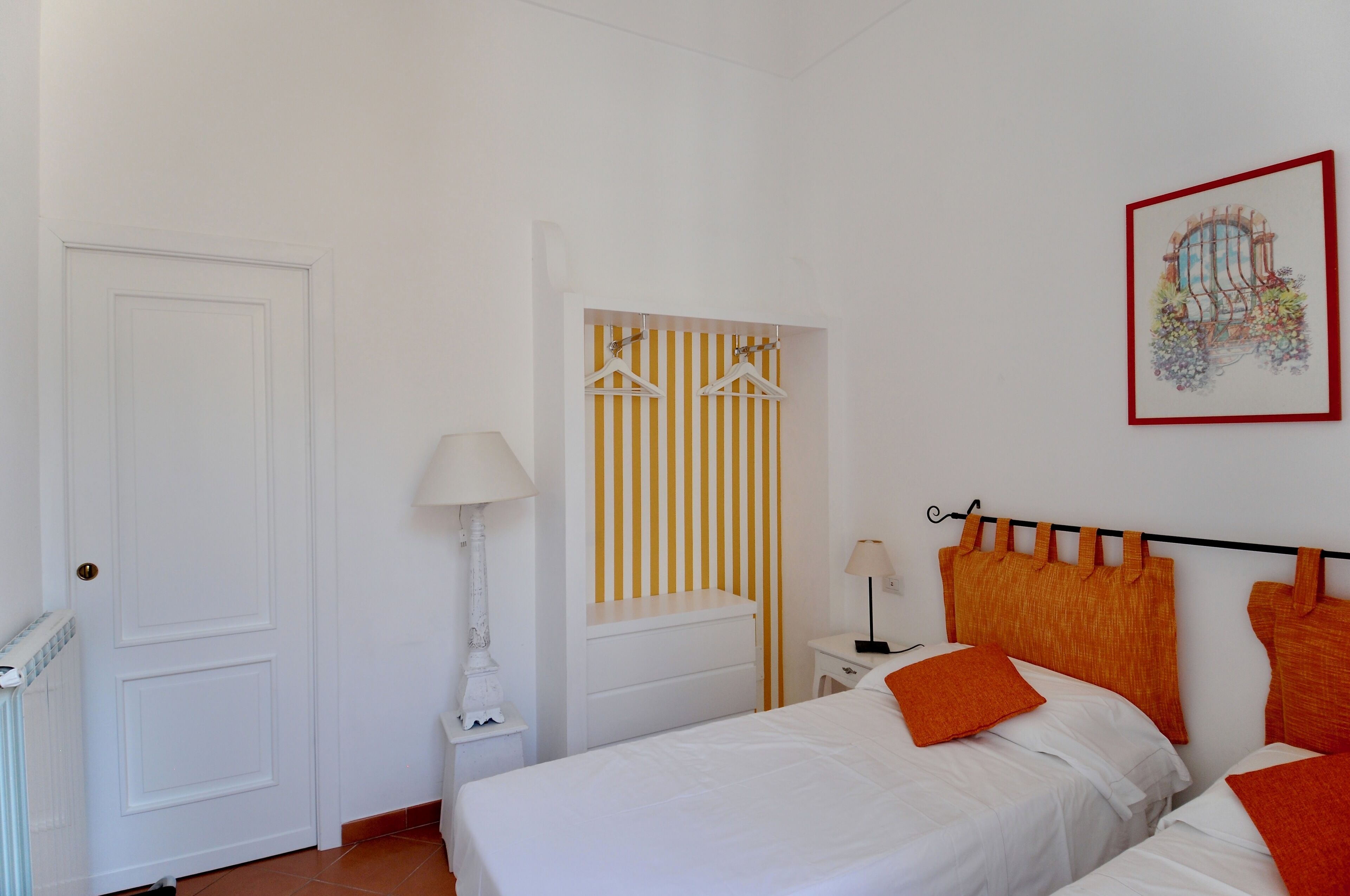 Une chambre avec deux lits, une porte blanche, une fenêtre avec des stores jaunes et un tableau encadré au mur.