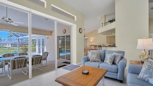 A spacious living area with a light blue sofa, a wooden coffee table, and a dining table with chairs. There is a sliding glass door leading to a balcony with outdoor furniture.
